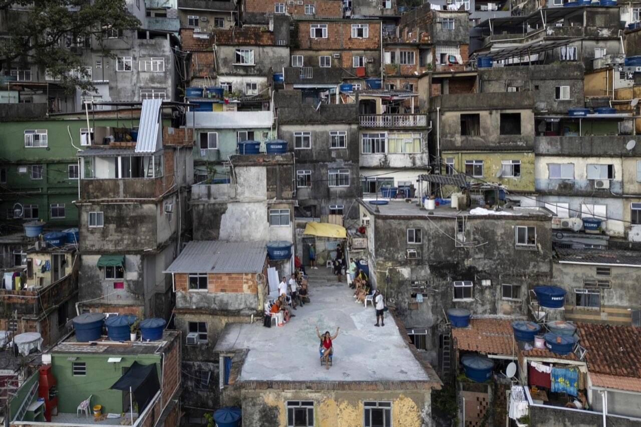 Drone-POV photo of a Rocinha rooftop and the surrounding favela — woman on a rooftop with arms raised, dense vertical favela architecture all around her.